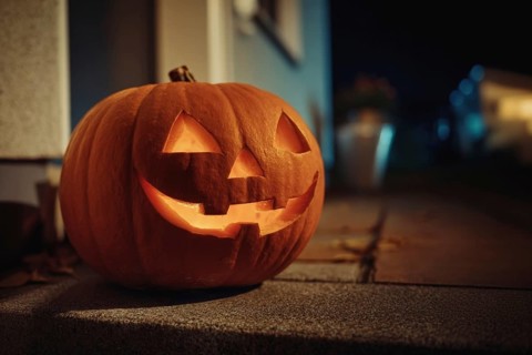 A carved, glowing pumpkin with a smiling face stands on a wall in front of a house at night,