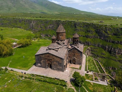 Historic stone church on a green plain in front of a mountain landscape under a blue sky, aerial