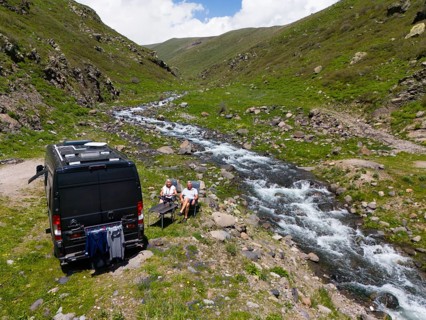 A campervan stands next to a river in the mountains where people sit at a table and enjoy nature,