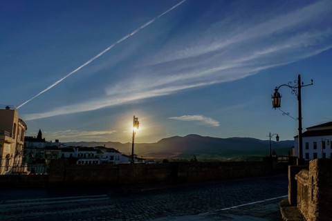 Sun rising behind mountains, illuminating street lamps on cobblestone bridge at dawn in ronda,