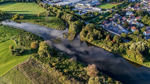An idyllic landscape shows a gently flowing river, surrounded by lush vegetation and a small town