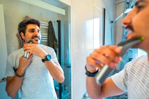 Young man trimming his beard using an electric razor, looking at himself in the bathroom mirror