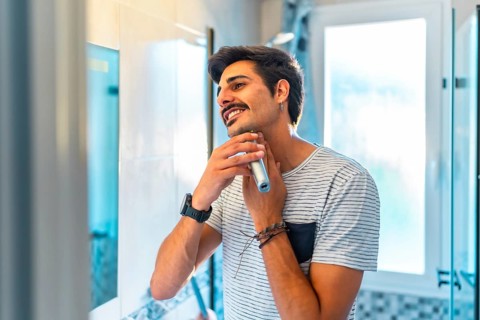 Happy young man trimming his beard in the bathroom mirror using an electric razor, enjoying his