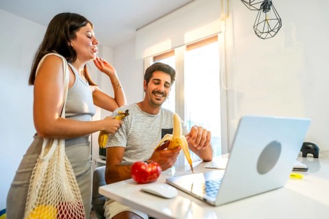 Young couple smiling and enjoying bananas while working on a laptop in a bright, modern home