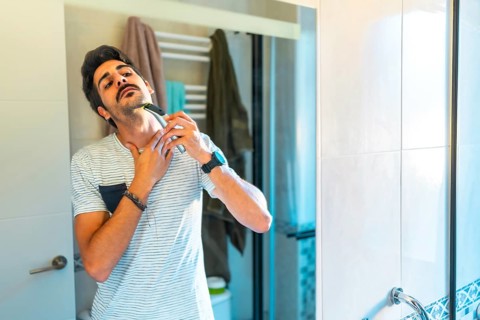 Young man carefully shaving his neck with an electric razor, looking in the bathroom mirror during