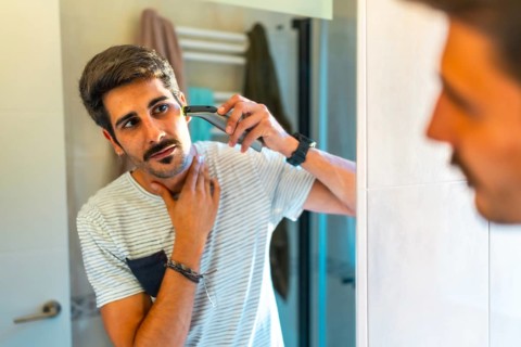 Concentrated young man trimming his beard with electric razor in bathroom mirror, morning routine,