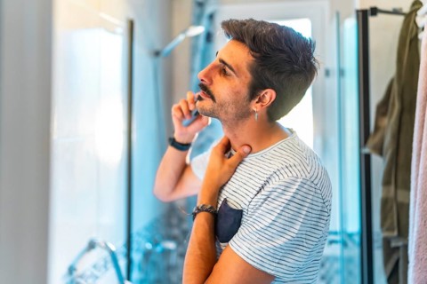 Young man trimming his mustache with an electric razor and touching his neck while looking in the