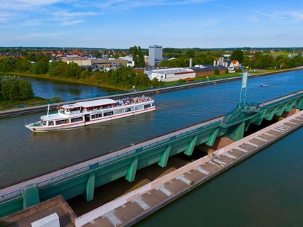 A passenger ship sails on a canal over a large bridge, surrounded by city buildings and nature,