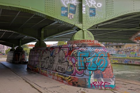 Under a bridge with graffiti on the retaining walls and a river in the background, Am Donaukanal