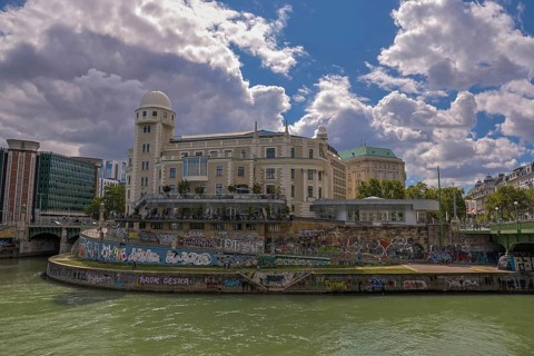 A historic building by the river with graffiti, under a clear sky, surrounded by urban surroundings
