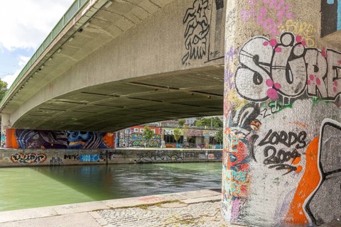 Under a bridge on the river with graffiti-covered columns and riverbank surroundings, Am Donaukanal