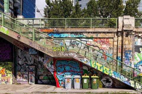 Various graffiti artworks on a concrete wall next to stairs and rubbish bins, Am Donaukanal Innere