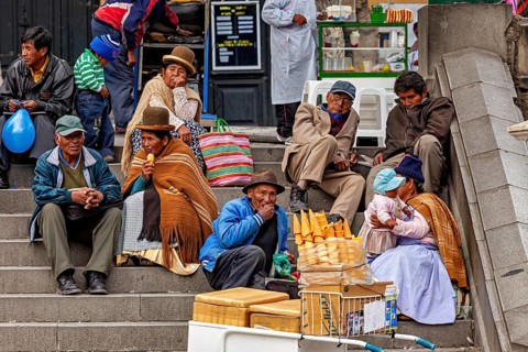People in traditional dress sit on a staircase and talk near a market stall with ice cream,
