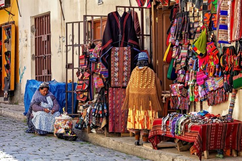 A woman sits in front of a market stall selling a variety of colourful textiles and traditional