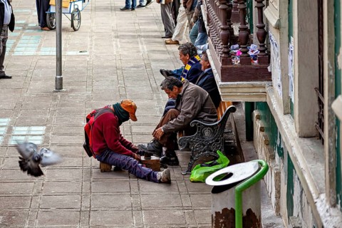 A shoeshine boy shines shoes on a street while passers-by and a pigeon are visible, shoeshine boy