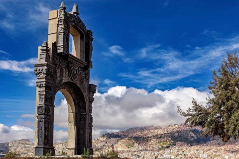 A stone gate with ornate decorations in front of a sprawling cityscape and a blue sky, The gate at