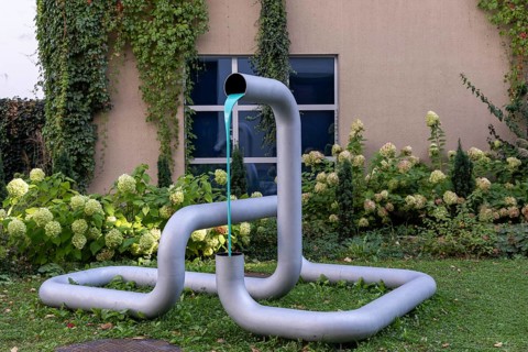 Metal sculpture in the garden with flowing coloured water and plants in the background, city centre