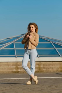 A young woman stands outdoors, wearing a light jacket and jeans. She holds her jacket gently while