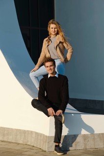 A young man sits on a low wall with his leg bent while a woman stands above him, both smiling. The
