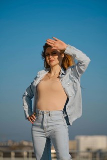 A woman stands outdoors on a sunny day, shielding her eyes from the sunlight. She wears a light
