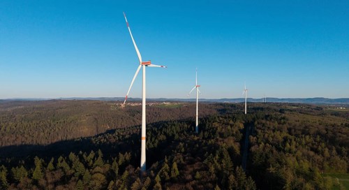 Several wind turbines tower above the treetops of a forest under a blue sky, near Schorndorf in the