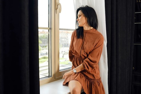 A woman in a brown dress relaxes by a large window, gazing thoughtfully outside. Sunlight filters