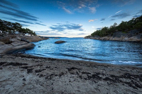 Small bay with sand and rocks, surrounded by pine trees under a partly cloudy sky, Skjeberg,