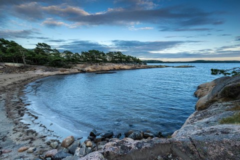 Small bay with sandy beach, surrounded by rocks and pine trees under a partly cloudy sky, Skjeberg,