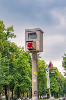 Traffic surveillance camera on a pole, surrounded by trees and road signs under a cloudy sky,