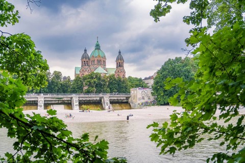 Imposing church behind a river, lined with trees and sandbanks under a cloudy sky, Munich, Germany