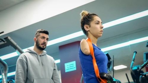 A young female athlete with limb loss trains in a fitness gym with the support of her coach,