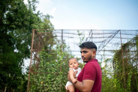 A man holds a baby outdoors by a metal structure with greenery in a relaxed setting-Dhaka,