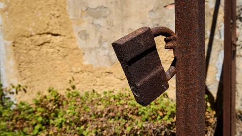 Rusted lock on a metal post in front of a weathered wall with plants in the background, Old silk