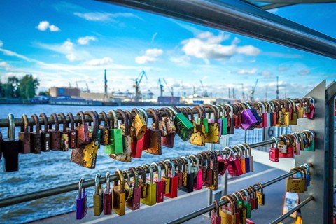 Many colourful love locks on a railing with a view of the harbour and cranes. View from the jetties