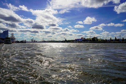 The Norderelbe with wavy water under a sunny sky, with urban background and silhouettes of cranes.