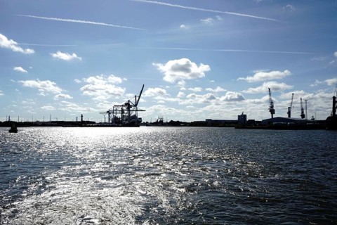 An industrial harbour with cranes and reflecting sunlight on the water under a blue sky. The view