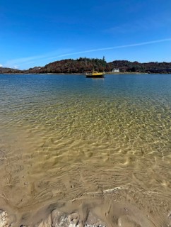 Clear water on a beach with a view of a yellow boat and wooded island in the background, Silver