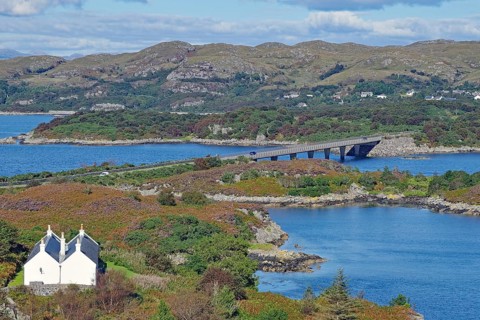A white house set in a picturesque landscape of bridge, loch and rolling hills under a blue sky,