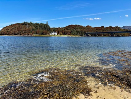 View over a loch with bridge and house surrounded by wooded hills under a blue sky, Silver Sands of