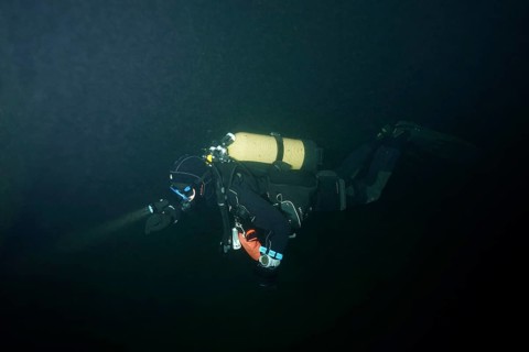 A diver in a diving suit explores a dark underwater environment. Dive site wreck of the Jura, Lake