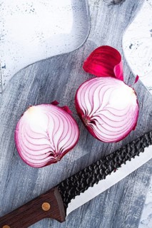 A vibrant red onion is cut in half, on a rustic wooden chopping board beside a sharp knife, ready