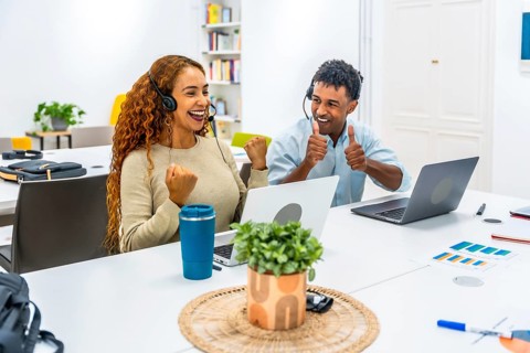 Two diverse call center agents wearing headsets and working on laptops, celebrating their