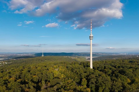 Television tower rises above a dense forest under a clear, slightly cloudy sky,