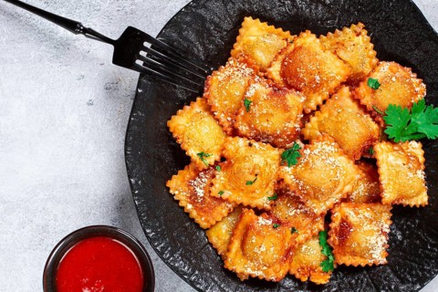 Fried ravioli with parmesan cheese, served with red sauce and garnished with herbs, on a black