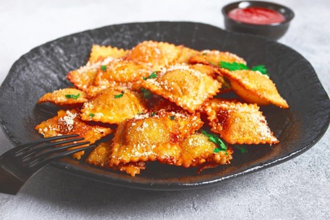 Fried ravioli with parmesan cheese, served with red sauce and garnished with herbs, on a black