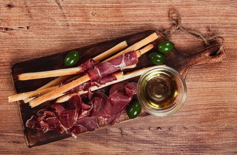 An appetizer of prosciutto, grissini and olives, served on a wooden board, with olive oil
