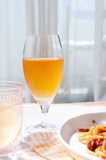 A glass filled with a light amber drink sits beside a plate of pasta. Bright sunlight streams
