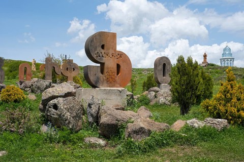 Stone sculptures in the shape of letters on a green meadow, surrounded by rocks and bushes under a