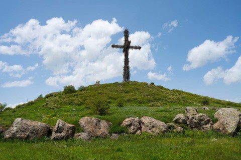 Metal cross on a green hill under a blue sky, surrounded by large stones, Monument to the Holy