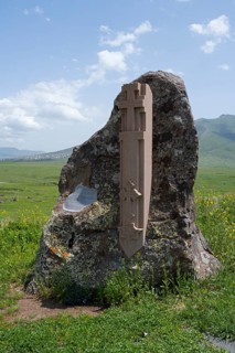 Stone with cross relief in front of a vast green landscape and blue sky, at the Monument of the
